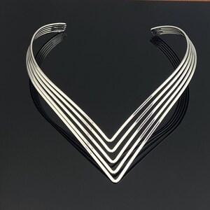 May include: A silver-toned metal collar necklace featuring five distinct, parallel V-shaped bands that converge into a sharp point at the center. The rigid, open-ended design creates a layered, geometric appearance with a smooth, polished finish.