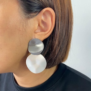 以下が含まれることがあります： シルバーカラーの2ピースディスクイヤリング。上のディスクは小さくマットで、下のディスクは大きくブラッシュ仕上げです。イヤリングは耳に装着され、黒いシャツを着ています。