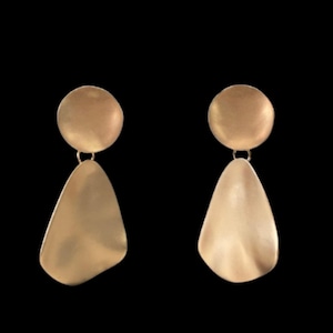 May include: A pair of gold-toned drop earrings featuring a circular top stud connected to a larger, irregular, organic-shaped pendant. The metal surface has a smooth, matte finish with soft, flowing curves that catch the light, creating a subtle glow.