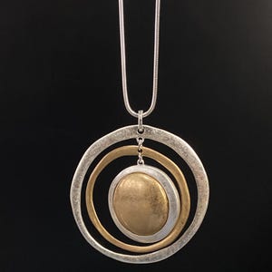Puede incluir: Un collar plateado con un colgante que presenta tres círculos concéntricos. El círculo exterior es plateado, el del medio es dorado y el círculo interior es un disco dorado. El collar cuelga de una cadena plateada.
