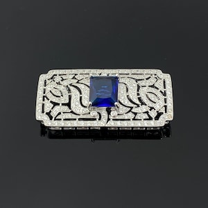 May include: A rectangular silver-toned brooch with a large, square, deep blue gemstone at its center. The brooch is encrusted with small, clear gemstones and features an openwork design with swirling patterns. The brooch is displayed on a reflective black surface.
