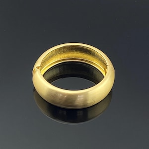 May include: A gold-toned hinged bangle bracelet with a smooth, brushed matte finish on the exterior surface. The circular jewelry piece features a visible hinge mechanism on the left side, allowing it to open and close securely around the wrist.