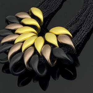 May include: A necklace featuring a cluster of teardrop-shaped elements in matte gold, bronze, and black finishes. The pendant is attached to multiple strands of fine black chain, creating a layered and textured appearance with a fan-like arrangement.