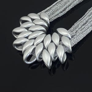 May include: A silver-toned necklace featuring a cluster of smooth, matte-finished, petal-shaped beads arranged in a fan-like formation. The central focal point is attached to multiple fine, delicate silver-toned metallic chains that extend outward.