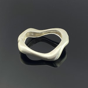 Könnte beinhalten: Ein silberfarbener Klapparmreif mit einer unregelmäßigen, organischen und wellenförmigen Silhouette. Die Metalloberfläche weist ein gebürstetes, mattes Finish auf, mit einem sichtbaren Scharniermechanismus auf der linken Seite, der eine fließende Form bildet.