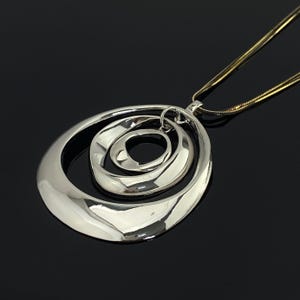 Könnte beinhalten: Eine silberfarbene Anhängerkette mit drei konzentrischen, unregelmäßig geformten ovalen Ringen mit polierter, reflektierender Oberfläche. Der Anhänger hängt an einer dünnen, flexiblen goldfarbenen Schlangenkette, die am größten Ring befestigt ist.