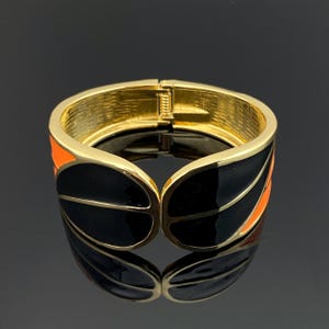 May include: A hinged gold-tone metal cuff bracelet featuring a bold design with glossy black enamel panels and vibrant orange accents. The interior of the bracelet has a textured finish, and the piece includes a secure metal clasp mechanism.