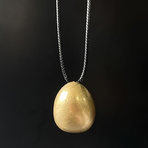 Könnte beinhalten: Ein goldfarbener Anhänger in Eiform hängt an einer schwarzen Kordelkette. Der Anhänger hat eine glatte, reflektierende Oberfläche. Die Halskette wird vor einem dunklen Hintergrund präsentiert, der die Farbe und Form des Anhängers hervorhebt.