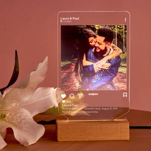 Op de afbeelding: Een gepersonaliseerde fotonachtlamp met een stel dat elkaar omhelst, weergegeven in een helder acryl frame. Het frame wordt verlicht door een LED-licht en rust op een houten voet. De tekst "Laura & Paul" en andere details in social media-stijl zijn zichtbaar.