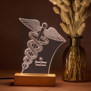 Può includere: Un simbolo medico illuminato con un design di caduceo, con un bastone intrecciato da due serpenti e sormontato da ali. Il simbolo è illuminato di bianco e poggia su una base di legno. Il testo "Dr. David Peterson, Orthopedic Surgeon" è visibile.
