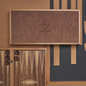 Könnte beinhalten: Ein Backgammon-Set mit einer braunen Leder-Spielfläche. Das Leder ist mit den Initialen "J.M." und dem Namen "John Miller" mit einem Datum geprägt. Holzspielsteine sind zu sehen.
