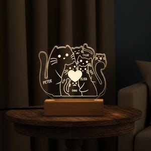 Op de afbeelding: Een gepersonaliseerd nachtlampje voor een kattenfamilie. Het acryllicht toont een familie van katten met namen gegraveerd op de voorkant. Het licht staat op een houten voet en wordt verlicht met een warme gloed. De namen zijn Peter, Olive, Emma, Elisa, Tylor en Kate.
