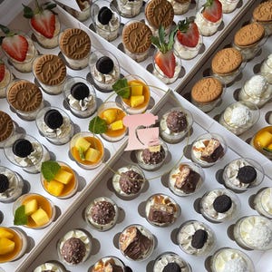 Puede incluir: Una variedad de mini postres en vasos transparentes, dispuestos en una caja blanca. Los postres incluyen fresas, galletas, chocolate y trozos de mango, ofreciendo una variedad de sabores y texturas. Una presentación visualmente atractiva para una fiesta.