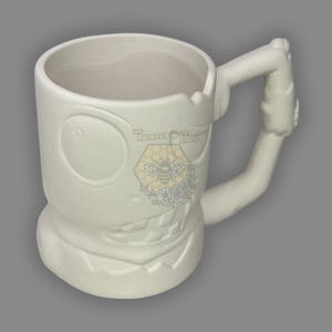 May include: A white ceramic mug with a cartoon monster design. The mug features a large eye, a toothy grin, and a handle shaped like an arm. The Painted Hive Studio logo is visible on the mug.