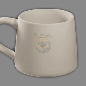 May include: A beige ceramic mug with a curved handle. The mug features a logo with a bee inside a honeycomb, surrounded by flowers, and the text "The Painted Hive Studio". The mug has a wide opening and a rounded base.