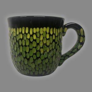 May include: A ceramic mug with a black rim and handle, and a green and yellow dotted pattern covering the body. The mug has a curved handle and a wide opening, suitable for hot or cold beverages.