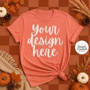 Op de afbeelding: Een koraalkleurig T-shirt met korte mouwen en de witte tekst "your design here" in cursief schrift in het midden. Het shirt heeft een ronde hals en ligt plat met een geknoopt detail aan de rechteronderkant van de zoom.