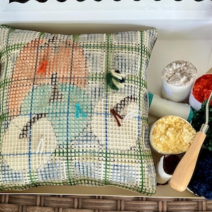 May include: A latch hook craft kit featuring a square canvas with a printed plaid pattern and a partially completed design of three circular shapes. The kit includes a wooden-handled latch hook tool and several small containers of colorful yarn.