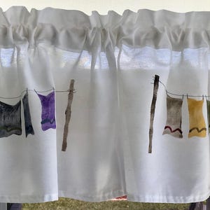 May include: White valance with a watercolor painting of clothes hanging on a clothesline. The clothes are painted in shades of yellow, orange, purple, blue, and green. The valance has a ruffled edge and is made of a lightweight fabric.