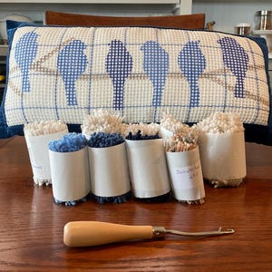 May include: A blue and white pillow with a cross-stitch design of birds on a branch. The pillow is on a wooden table with seven rolls of yarn in various colors, including white, beige, and blue. A wooden crochet hook is also on the table.