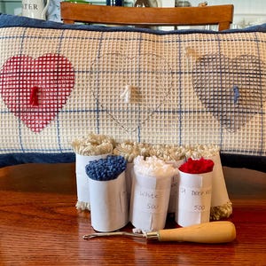 May include: A handmade pillow with a blue denim border and a grid pattern. The pillow features three heart designs, one red and two blue. Several rolls of yarn in white, blue, and red are in front of the pillow, along with a wooden-handled rug hook.