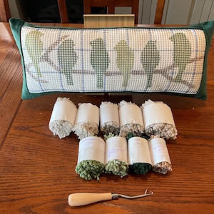 May include: A green and white needlepoint pillow with a design of birds on a branch. The pillow is on a wooden table with several skeins of yarn and a needle tool.