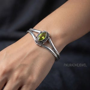 Könnte beinhalten: Ein silberner Armreif mit einem marquise-geschliffenen grünen Edelstein in der Mitte. Das Metallband ist in zwei parallele Linien geteilt, die an der Steinfassung zusammenlaufen, welche mit kleinen Silberperlen und gedrehtem Draht verziert ist.