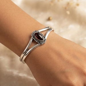 Può includere: Un bracciale rigido in argento con design a fascia divisa e una pietra preziosa rosso scuro a forma di marquise. La pietra è incastonata in una cornice d'argento testurizzata e decorata con piccole perline, creando un punto focale dettagliato.
