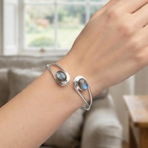 Puede incluir: Un brazalete de plata con dos extremos abiertos en forma de lágrima, cada uno engastado con una piedra preciosa de labradorita de corte ovalado y liso que muestra tonos azules y grises iridiscentes. La banda tiene un acabado pulido.