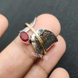Puede incluir: Un anillo de plata con diseño de hoja y una banda dorada. El anillo presenta una piedra preciosa ovalada roja. El anillo está en el dedo de una persona. El texto "PIKURADHEJEWEL" es visible.