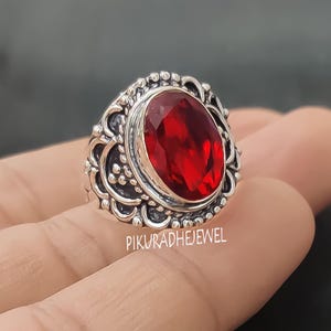Könnte beinhalten: Ein silberner Ring mit einem ovalen, tiefroten Edelstein. Der Ring hat ein verziertes, detailliertes Design um den Edelstein herum, mit einem strukturierten Band. Der Ring wird auf einer Hand vor dunklem Hintergrund gezeigt. Der Text "PIKURADHEJEWEL" ist sichtbar.