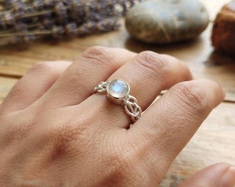 May include: A silver ring featuring a round, iridescent moonstone cabochon set in a bezel. The band is constructed from intricate, woven silver wire links that create a textured, openwork pattern around the finger, showcasing a polished finish.