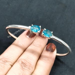 Puede incluir: Un brazalete plateado con dos gemas ovales de color azul brillante. El brazalete tiene un diseño sencillo y abierto, con las gemas muy juntas. Las gemas son de un color vibrante y llamativo, y el brazalete se muestra sobre un fondo oscuro.