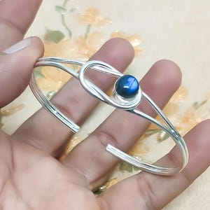 Pode incluir: Uma pulseira de punho prateada com uma pedra preciosa azul. A pulseira apresenta um design minimalista com um detalhe central em forma de nó que segura a pedra preciosa. O design aberto facilita o uso. A pedra preciosa é de um azul profundo.