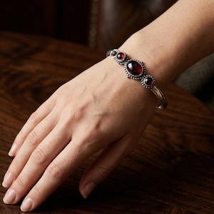 Può includere: Un bracciale rigido in argento con tre pietre preziose ovali rosso scuro incastonate in una lavorazione metallica decorata. La pietra centrale è la più grande, affiancata da due pietre più piccole, fissate in una cornice d'argento perlata.