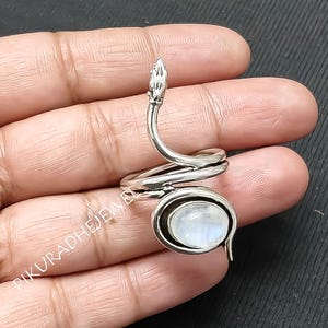 May include: A silver-toned ring in the shape of a snake, coiled around a central oval-shaped, iridescent moonstone. The snake's head is raised, and the ring is displayed on a hand.
