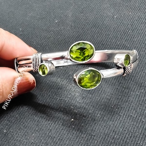 Op de afbeelding: Een zilverkleurige armband met vier ovaal geslepen, lichtgroene edelstenen. De armband heeft een uniek ontwerp met de edelstenen op verschillende punten. De achtergrond is donkergrijs.