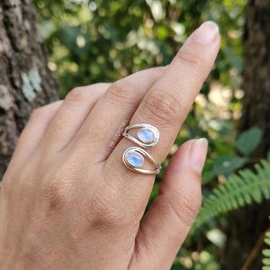 May include: A silver bypass ring featuring two oval-shaped moonstones with a blue iridescent sheen. The band is open and adjustable, showcasing a smooth, polished metal finish. The text PIKURADJEWEL is visible on the metal surface.
