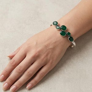 Könnte beinhalten: Ein silbernes Armband mit einer Anordnung von sechs ovalen, tiefgrünen facettierten Edelsteinen, die versetzt auf der Oberseite platziert sind. Das Metallband ist glatt und poliert und bildet einen klaren Kontrast zu den dunklen Steinen.