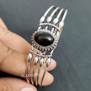 Peut inclure: Bracelet manchette argenté avec une pierre noire ovale au centre. Le bracelet présente un design détaillé avec des éléments décoratifs et des bandes parallèles. Le bijou est tenu par une main.