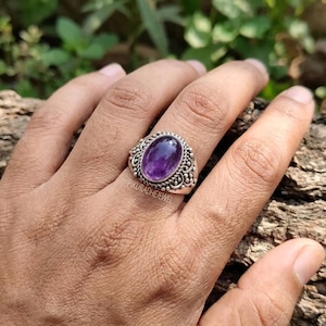 Op de afbeelding: Een zilveren ring met een centrale ovaalvormige paarse amethist edelsteen. De band is vervaardigd met ingewikkeld metaalwerk, inclusief kleine kraaldetails en wervelende patronen rondom de steen. De tekst PIKURADHEJEWEL is zichtbaar.