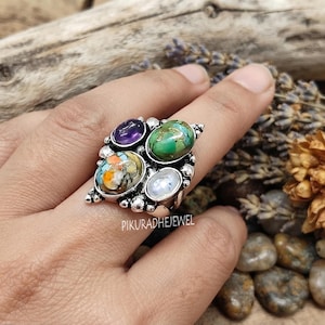 Op de afbeelding: Een zilveren ring met vier verschillende ovaalvormige edelstenen: een paarse amethist, een groene steen in turkooisstijl, een veelkleurige gespikkelde steen met oranje en blauw, en een doorschijnende witte steen in een decoratieve metalen zetting.