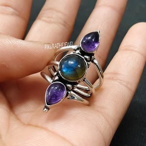 May include: A silver ring featuring a central round labradorite stone with blue and green iridescence, flanked by two teardrop-shaped amethyst stones. The ring has a multi-band design and the text "PIKURADHEJEWEL".