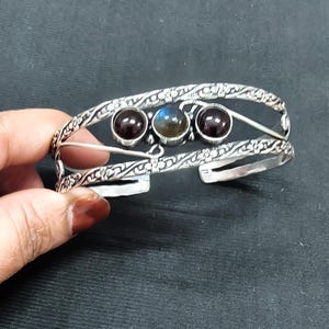 Op de afbeelding: Een zilveren manchetarmband met een ingewikkelde bloemmotiefband, voorzien van twee donkerrode ronde edelstenen aan de zijkanten en een centrale ronde iriserende blauwgrijze edelsteen. Het metaal heeft een textuur en geoxideerde afwerking.