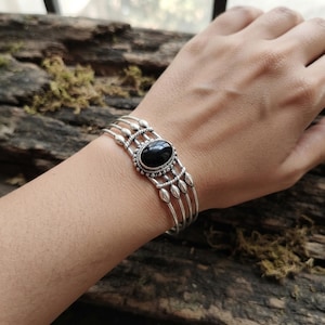 Könnte beinhalten: Ein silbernes Armband mit einem zentralen, ovalen schwarzen Stein in einer dekorativen Perlenfassung. Das Armband besteht aus vier dünnen, parallelen Silberdrähten, die an den Seiten zusammenlaufen und mit kleinen, glatten Silberperlen verziert sind.