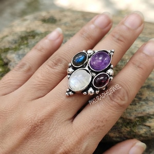 Può includere: Un anello in argento sterling con quattro pietre preziose ovali: una labradorite blu, un'ametista viola, una pietra di luna bianca e un granato rosso scuro. La montatura in metallo è decorata con piccole sfere. Il testo PIKURADHEJEWEL è visibile.