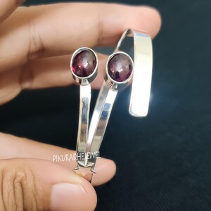 Peut inclure: Un bracelet manchette ouvert en argent orné de deux pierres précieuses grenat rouge foncé ovales et polies, serties dans des chatons lisses. La bande métallique est brillante et présente une petite étoile à la base. Le texte PIKURADHEJEWEL est visible.
