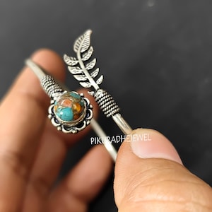 Könnte beinhalten: Ein silberfarbenes Armreif-Armband mit einem türkis-orangenen Stein in einer dekorativen Lünette. Das Armband hat ein Blattdesign und verdrehte Metallakzente. Der Text "PIKURADHEJEWEL" ist sichtbar.