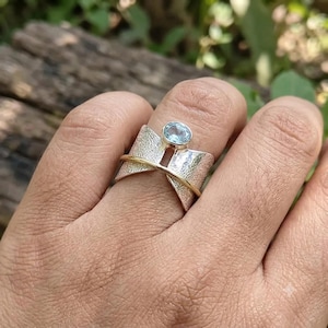 Op de afbeelding: Een brede zilveren ring met een getextureerde, gehamerde afwerking en een dunne, gladde goudkleurige band die aan de voorzijde kruist. De ring is in het midden voorzien van een kleine, ovale lichtblauwe edelsteen in een gepolijste zilveren zetting.