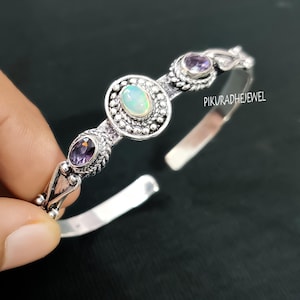 Puede incluir: Una pulsera de plata con un ópalo ovalado en el centro, flanqueado por dos amatistas ovaladas. La pulsera tiene un diseño detallado y ornamentado con el texto "PIKURADHEJEWEL".
